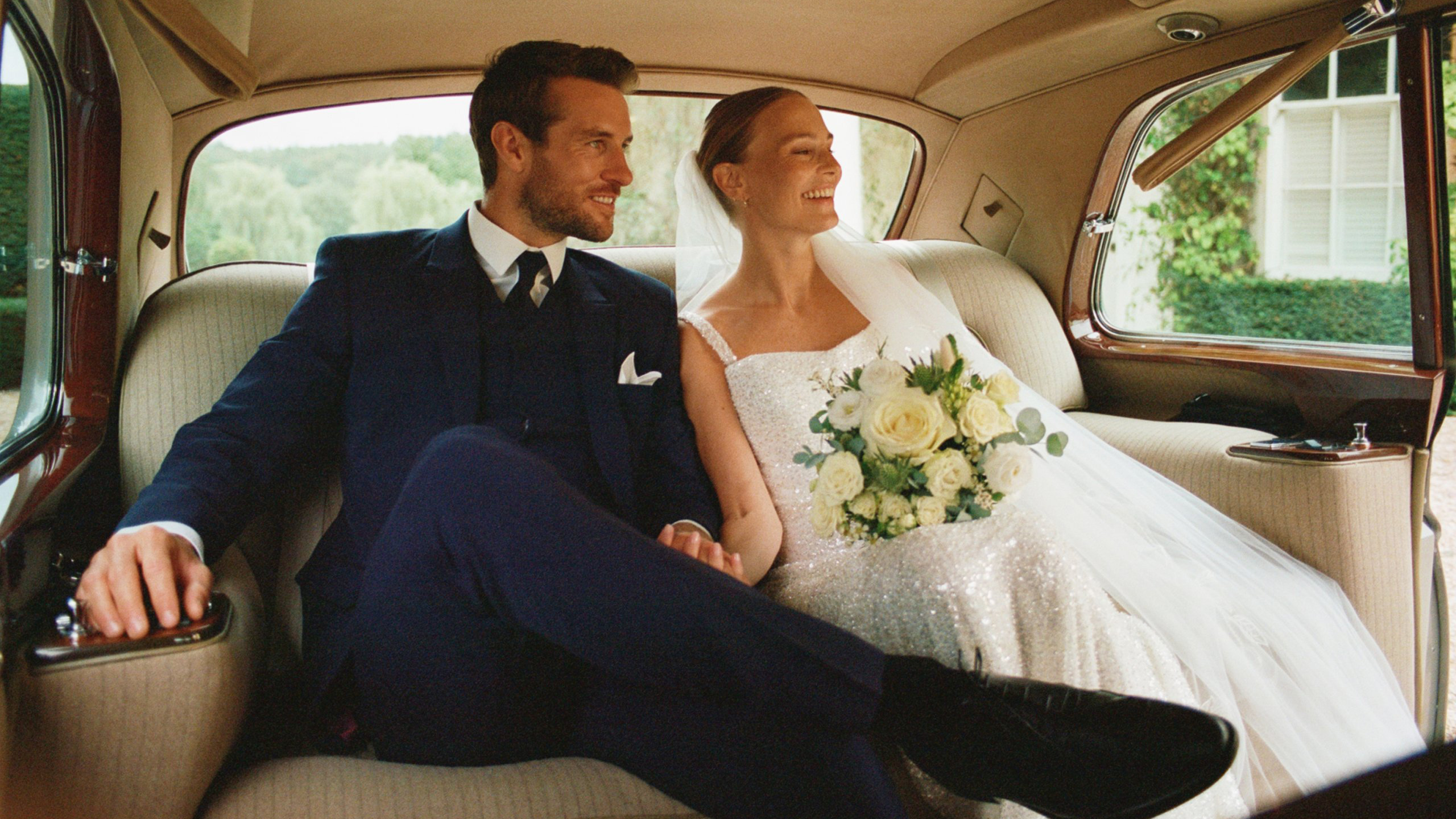Bride and groom seated in the rear of a classic Rolls-Royce wedding car smiling together.