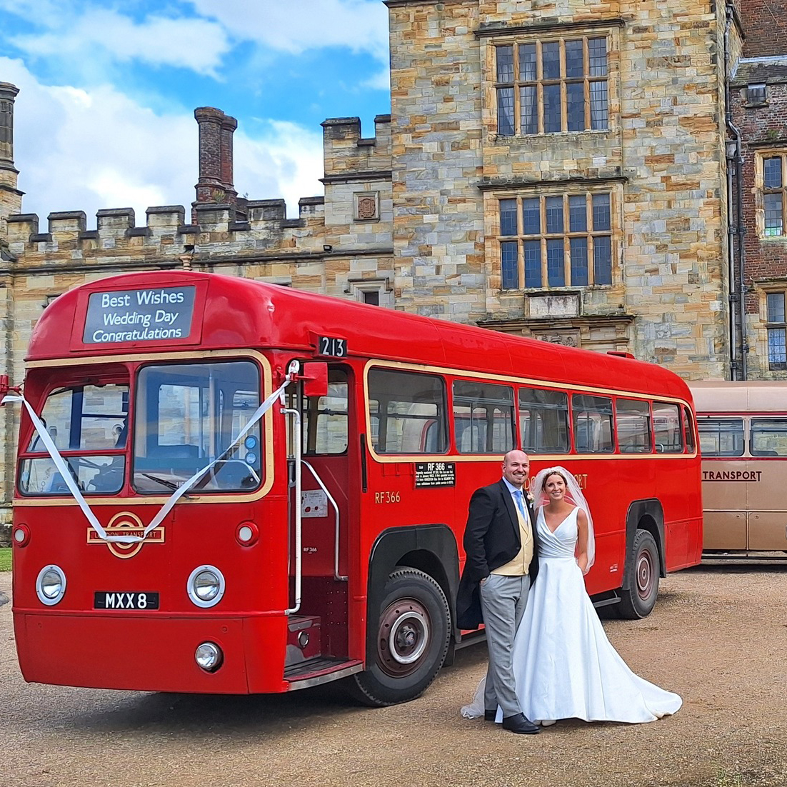 Bride wearing a white wedding dress standing beside her groom in front of a red and single decker vintage bus. A second gold single decker bus can be seen in the background with a large wedding venue in the far background.