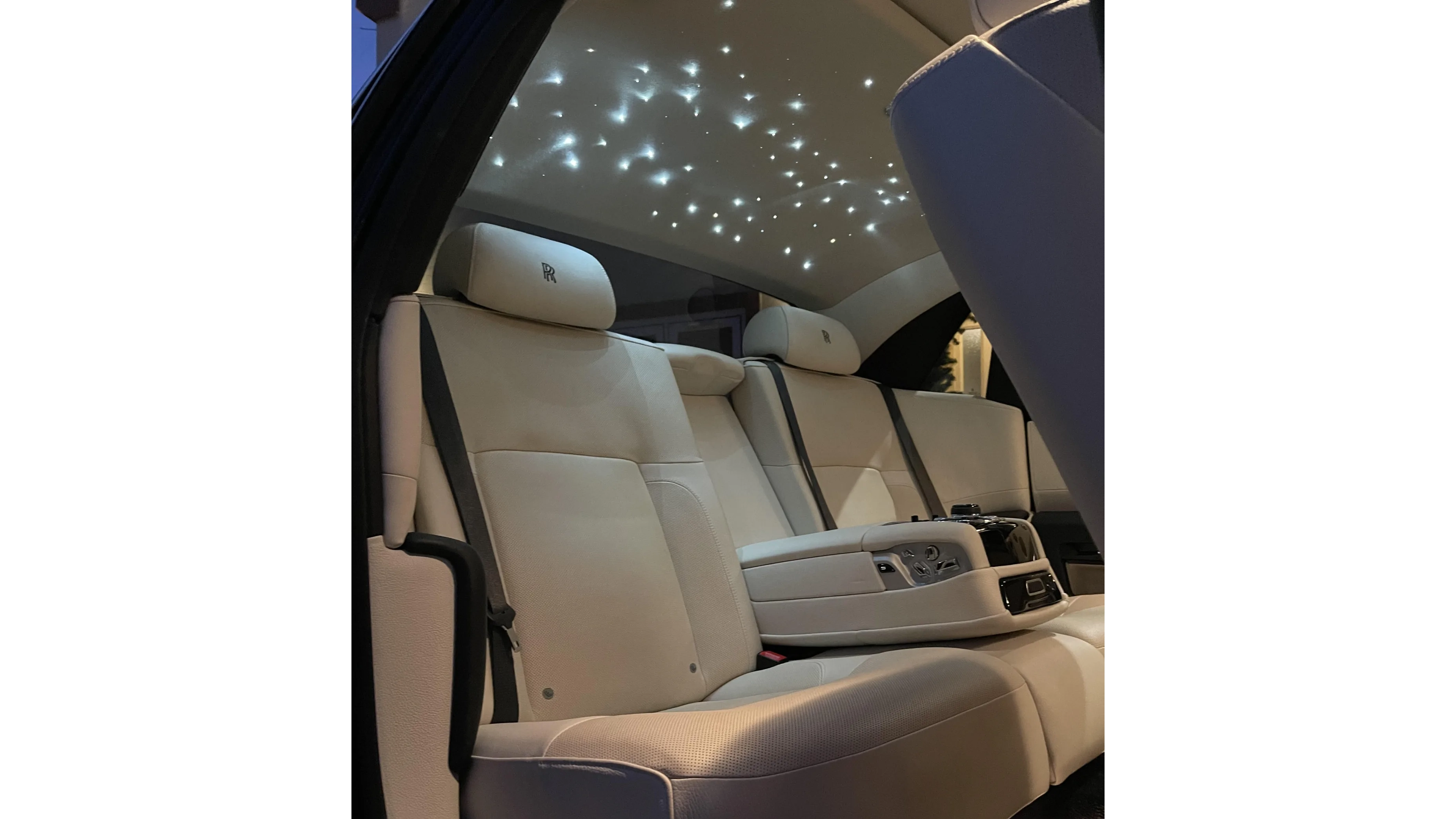 Rear interior of a Rolls-Royce Ghost showing tan leather seats and the illuminated starlight-style roof, photographed at night from the back seat.