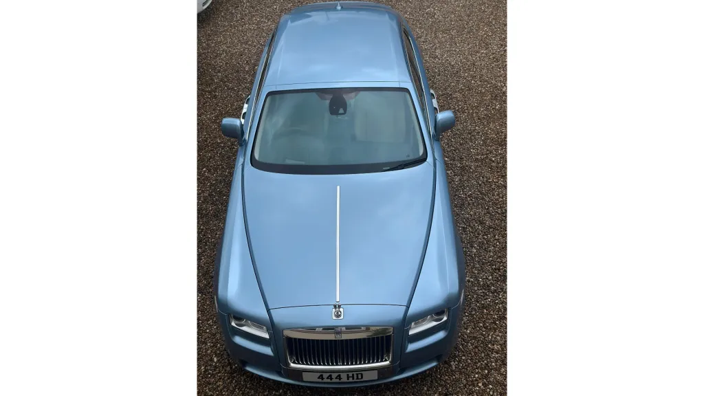 Aerial view of a light blue Rolls-Royce Ghost parked on gravel, showing its sleek bonnet, chrome details and roof.
