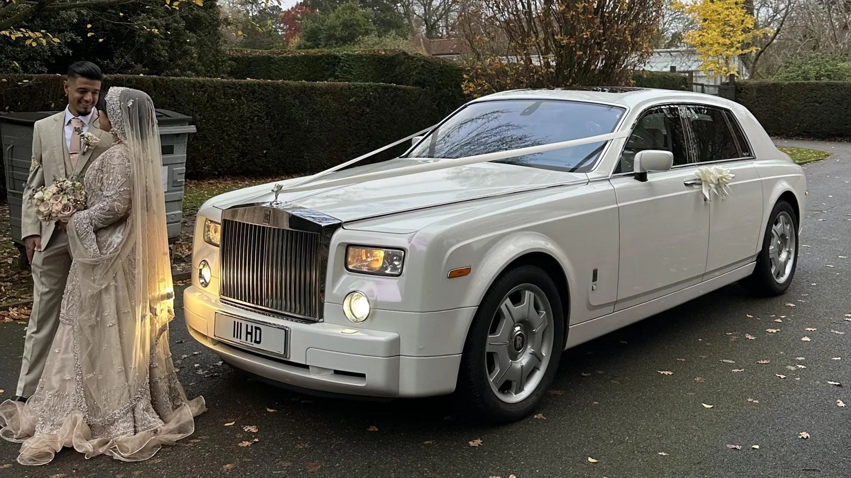 Asian couple standing beside a white Rolls-Royce Phantom, photographed at an outdoor wedding venue with soft golden light.