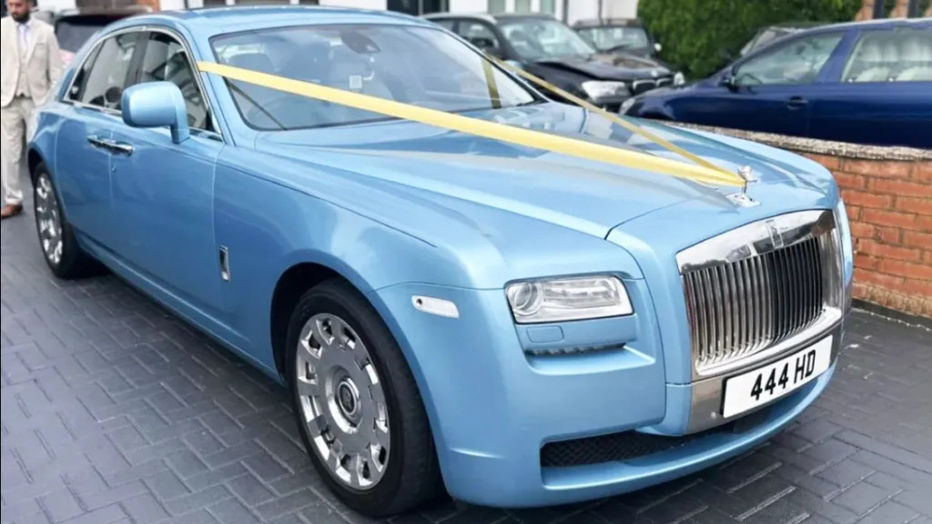 Front-angled view of a light blue Rolls-Royce Ghost decorated with yellow wedding ribbons, parked outside a brick building.