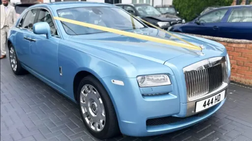 Front-angled view of a light blue Rolls-Royce Ghost decorated with yellow wedding ribbons, parked outside a brick building.