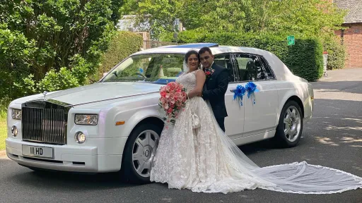 Bride and groom posing in front of a white Rolls-Royce Phantom decorated with blue and white wedding ribbons, parked on a tree-lined driveway.