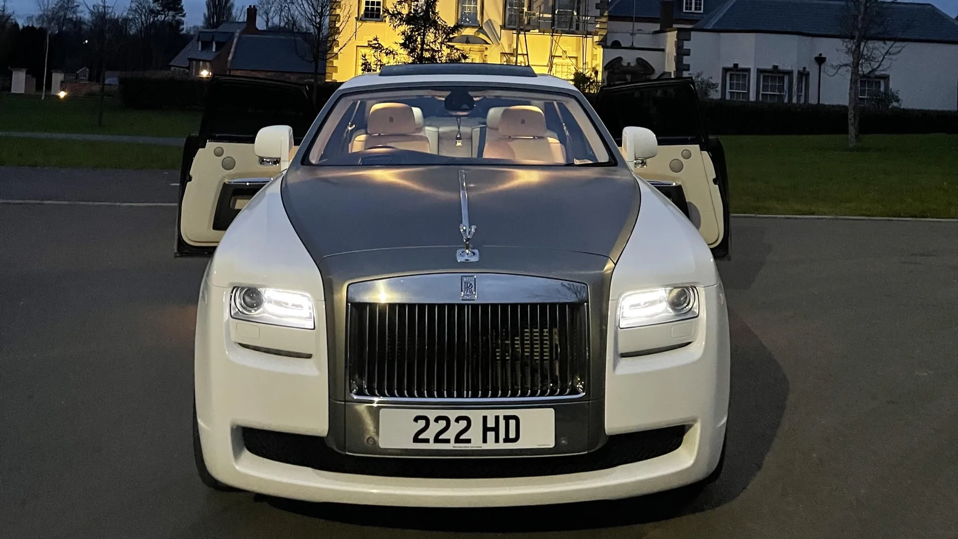 Front view of a white Rolls-Royce Ghost with a silver bonnet and both coach doors open, parked outdoors at dusk with soft evening light behind it.