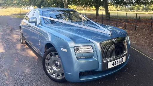 Side-front view of a light blue Rolls-Royce Ghost decorated with white wedding ribbons, parked near a grassy park area.