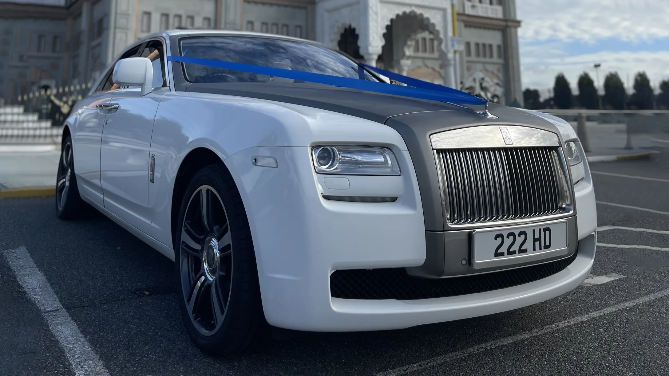 White Rolls-Royce Ghost with a silver bonnet decorated with royale blue wedding ribbons, parked outside a venue in natural daylight.