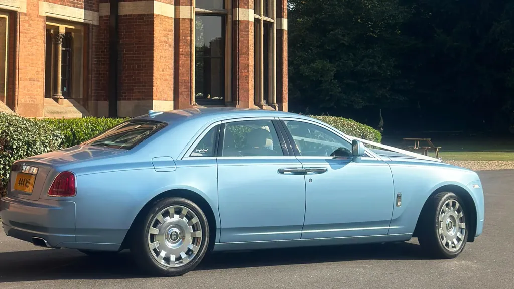 Right-side profile of a light blue Rolls-Royce Ghost parked on a driveway beside a brick house, showing its long elegant shape and silver alloy wheels.