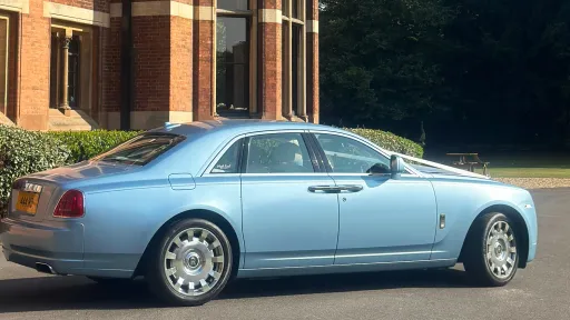 Right-side profile of a light blue Rolls-Royce Ghost parked on a driveway beside a brick house, showing its long elegant shape and silver alloy wheels.