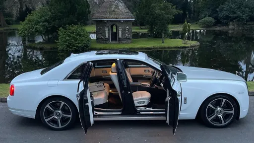 Side view of a white Rolls-Royce Ghost with both coach doors open, showing the tan interior and a landscaped garden in the background.