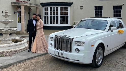 Bride and groom standing beside a white Rolls-Royce Phantom with subtle wedding ribbons, photographed outside a grand venue with stone pillars.