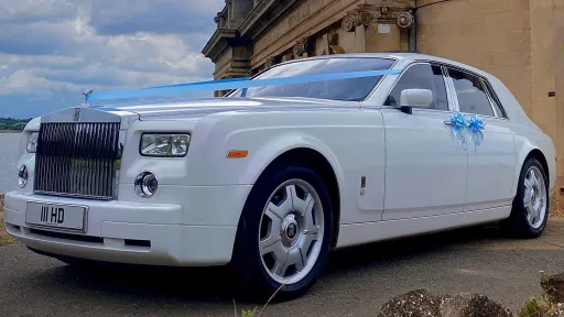 Side view of a white Rolls-Royce Phantom with blue wedding ribbons parked on a grassy drive, showcasing its chrome detailing and large alloy wheels.