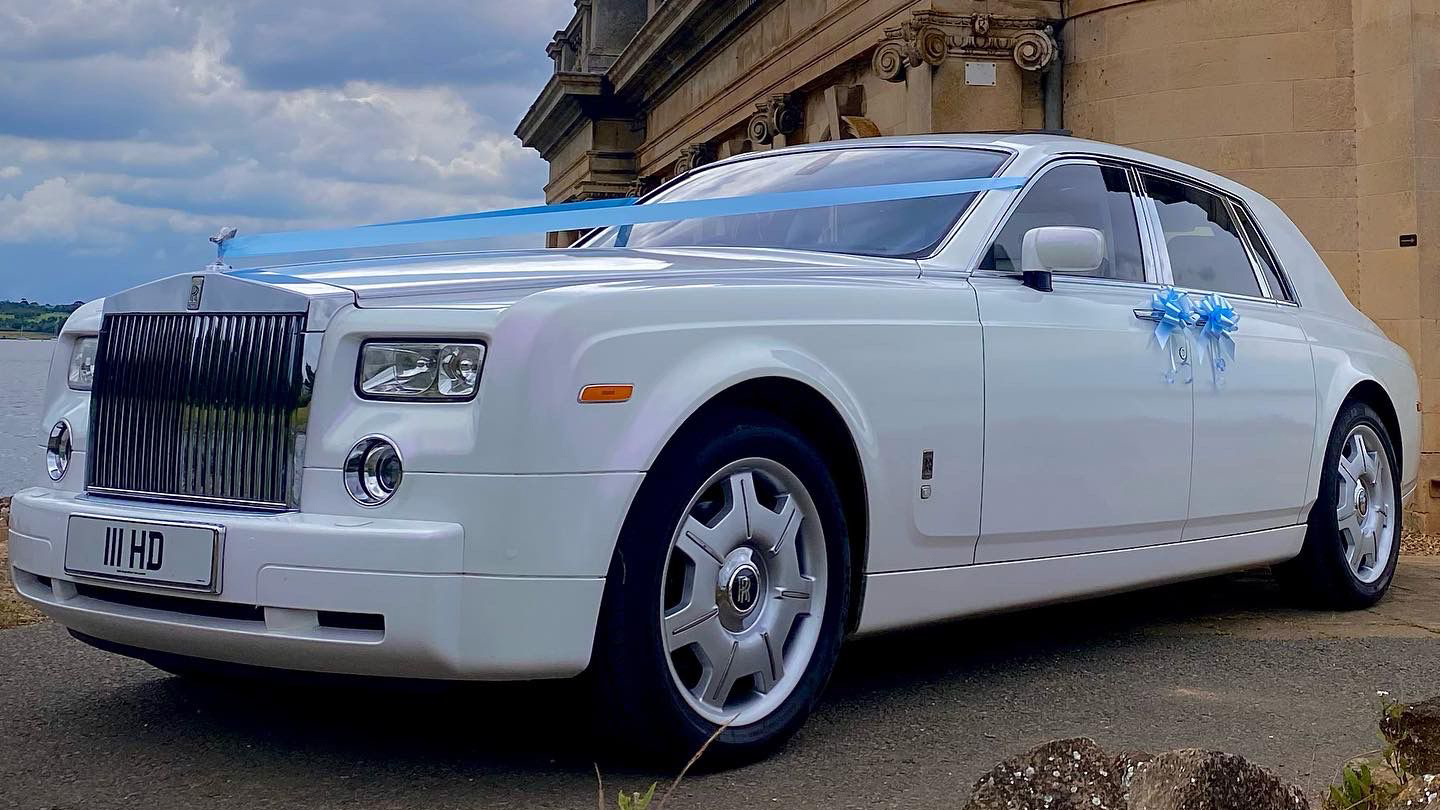 Side view of a white Rolls-Royce Phantom with blue wedding ribbons parked on a grassy drive, showcasing its chrome detailing and large alloy wheels.