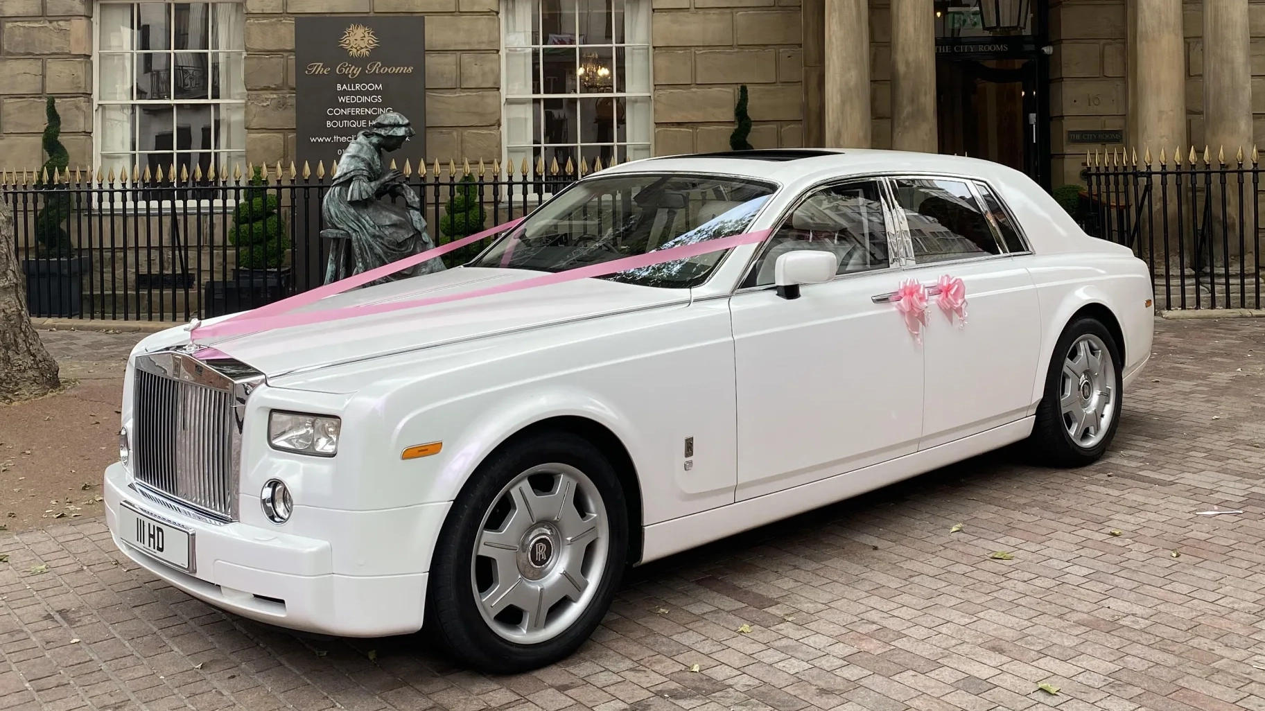 White Rolls-Royce Phantom decorated with pink wedding ribbons, parked outside a venue with brick pillar