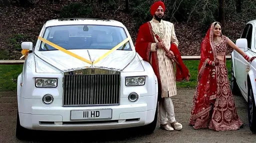 Front view of a white Rolls-Royce Phantom decorated with gold wedding ribbons, with an Asian couple posing in colourful outfits.