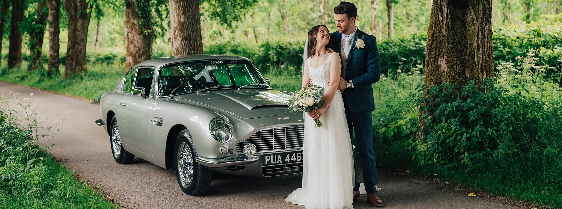Classic Aston Martin DB6 in silver parked in the middle of a path with greenery and trees on both side. Both Bride wearing a white dress is standing in front of the car with her groom.
