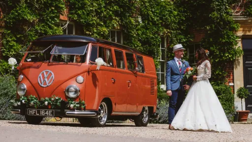 Volkswagen Splitscreen Campervan in Terracota colour with Bride and groom standing beside the vehicle. Vehicle is parked at a wedding venue in Essex