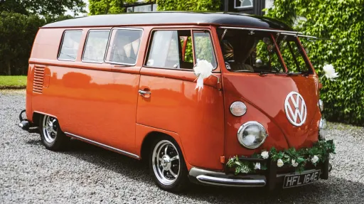 Right side viee of a terracota coral coloured classic Volkswagen Splitscreen Campervan dressed with wedding flowers on its front chrome bumper and white bows on wing mirror