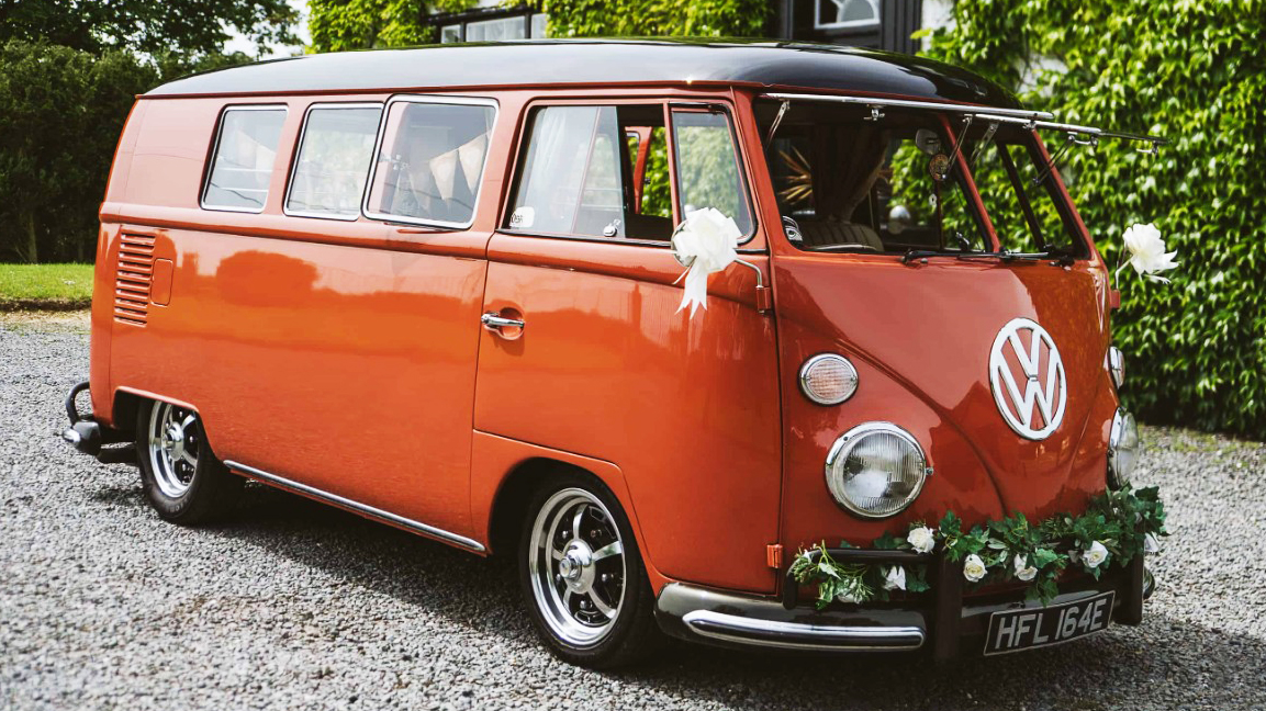 Right side viee of a terracota coral coloured classic Volkswagen Splitscreen Campervan dressed with wedding flowers on its front chrome bumper and white bows on wing mirror