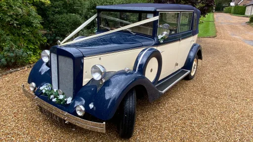 Cream and royal blue six-seater Regent convertible wedding car photographed from the front on a gravel driveway with greenery in the background.