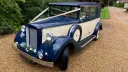 Cream and royal blue six-seater Regent convertible wedding car photographed from the front on a gravel driveway with greenery in the background.