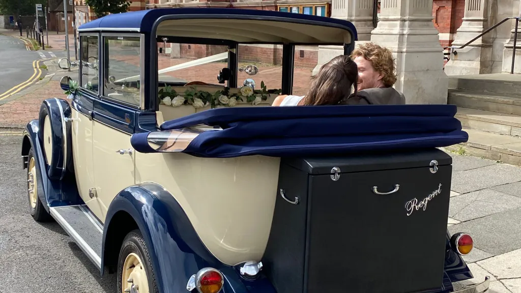 Bride and groom seated in the back of a cream and blue Regent convertible with the roof folded down, driving along a town street.