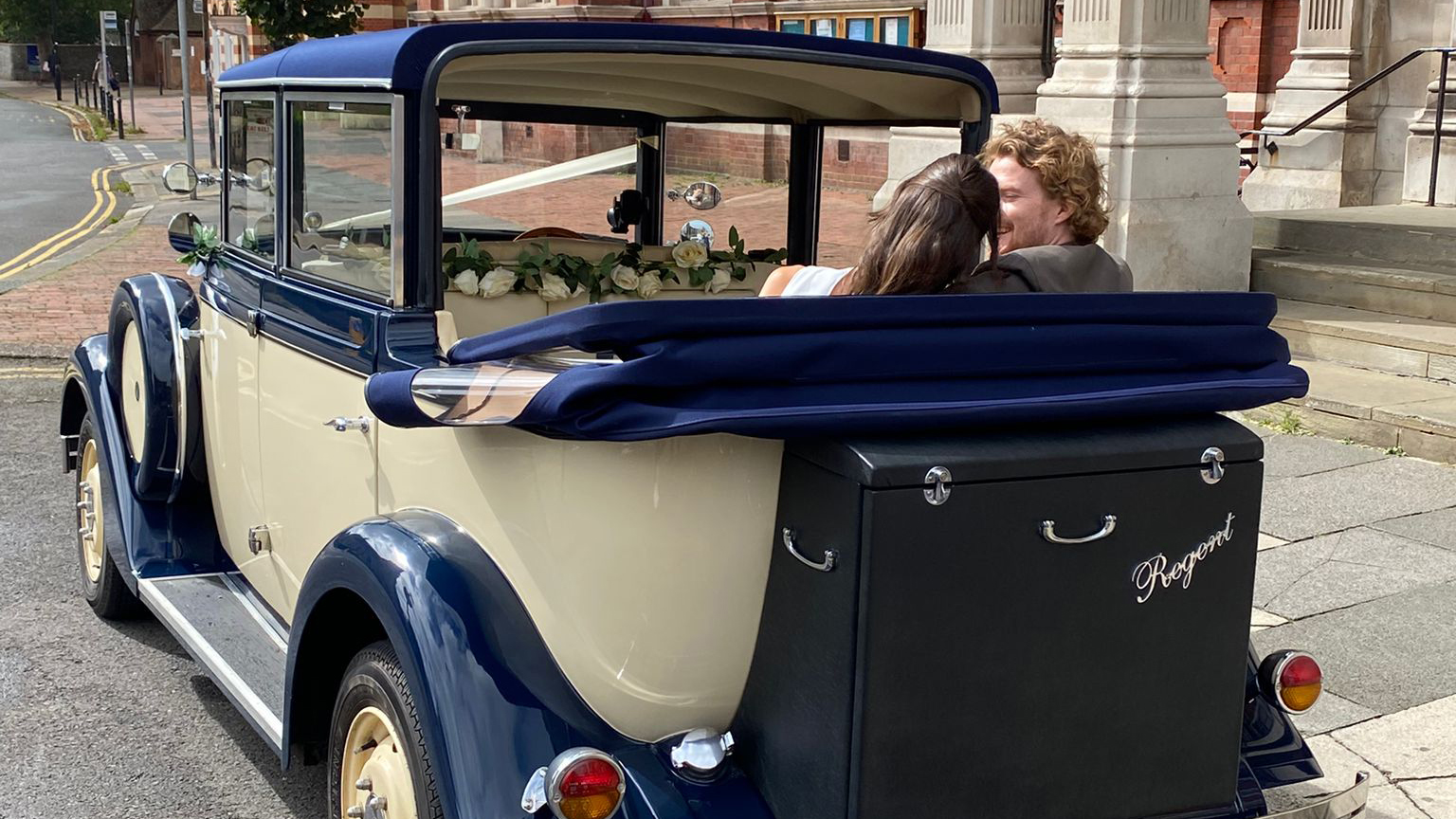 Bride and groom seated in the back of a cream and blue Regent convertible with the roof folded down, driving along a town street.