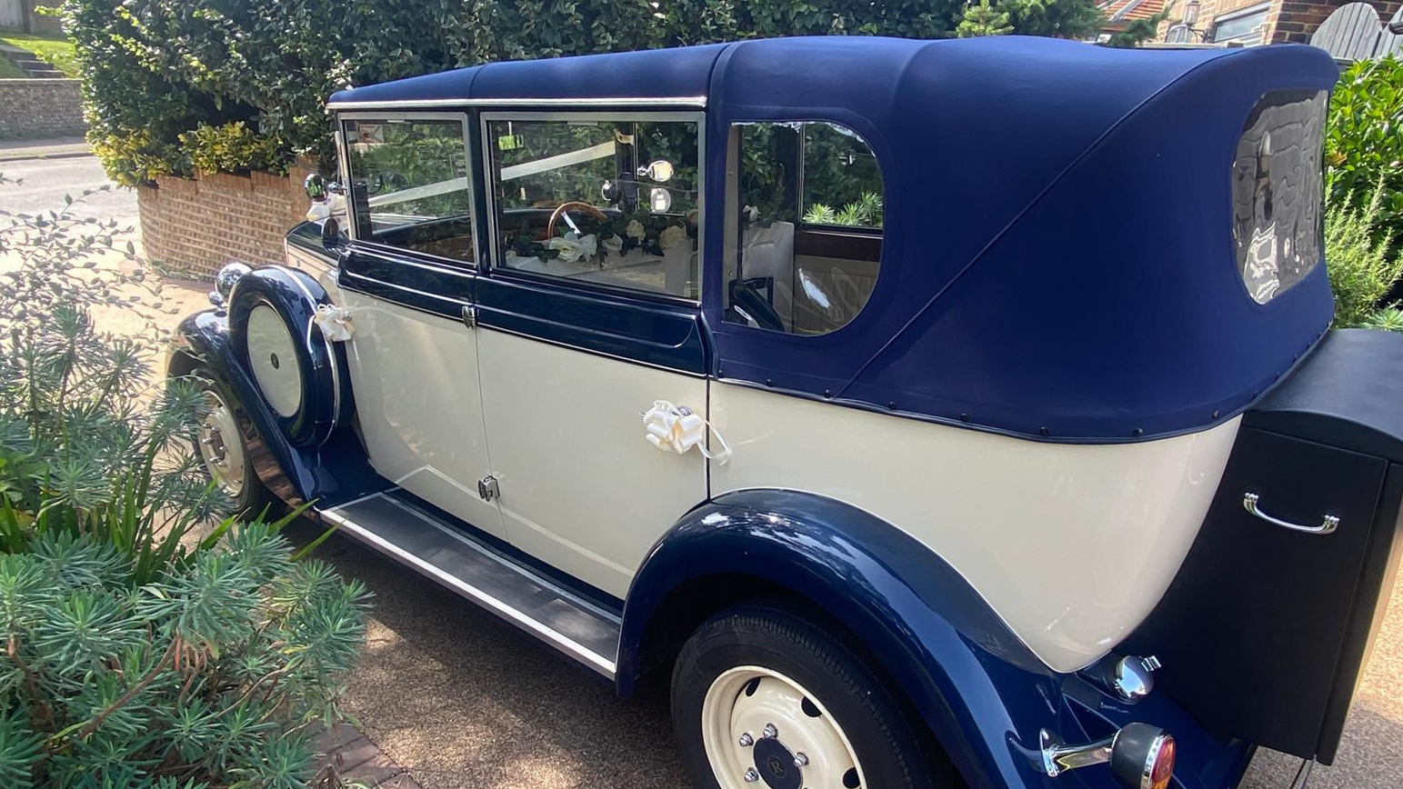 Side view of the cream and royal blue Regent vintage wedding car parked on a gravel driveway surrounded by greenery with his blue soft top roof closed and a black picnic trunk at the rear