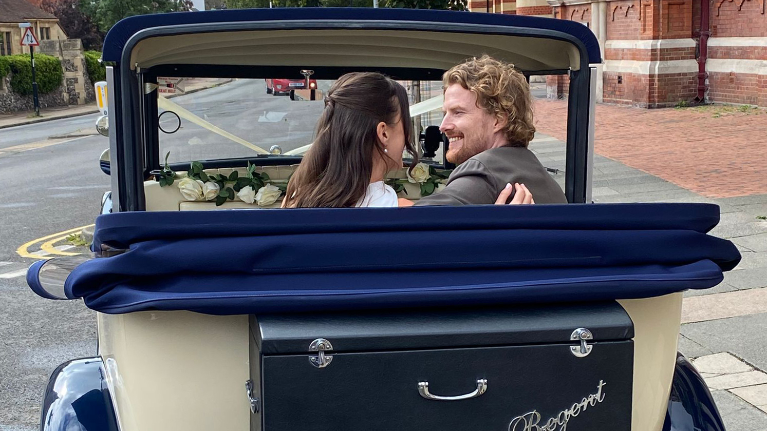 Rear view of a newly-married couple smiling together in the back seat of the Regent convertible with the navy roof folded down.