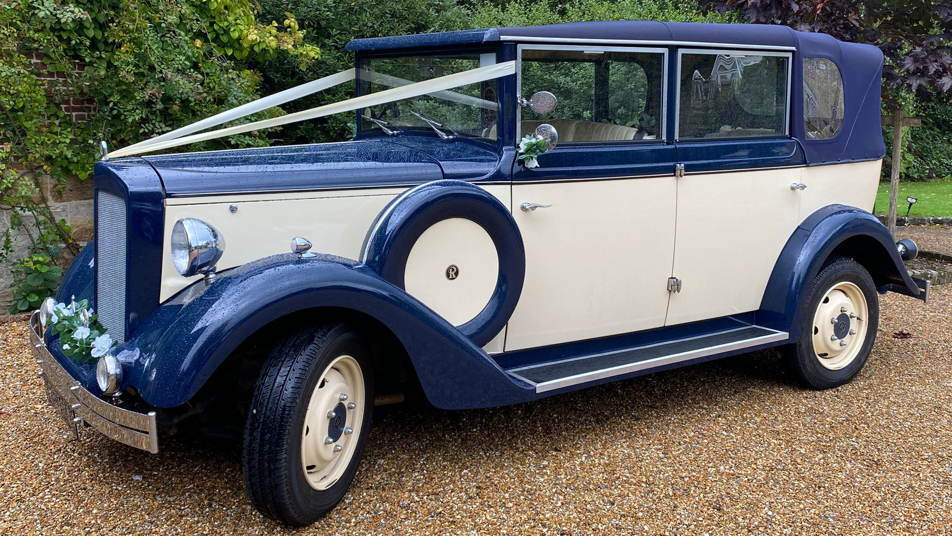 Cream and royal blue Regent wedding car photographed at an angle on a gravel driveway surrounded by trees and shrubs. Decorated with Wedding ribbons and sided mounted spare wheel.