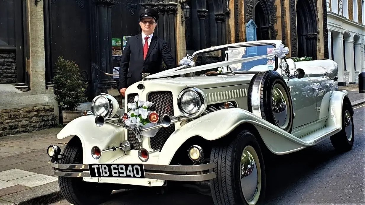 White Beauford vintage car decorated with white ribbons and bows and a fully uniformed chauffeur standing in the background wearing a Red tie and hat.