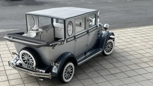 A silver Imperial Landaulette wedding car photographed from an elevated angle with the rear soft-top roof folded down, revealing the cream leather interior. The car sits on a paved driveway with subtle reflections on its grey bodywork and white-spoked wheels visible on both sides.