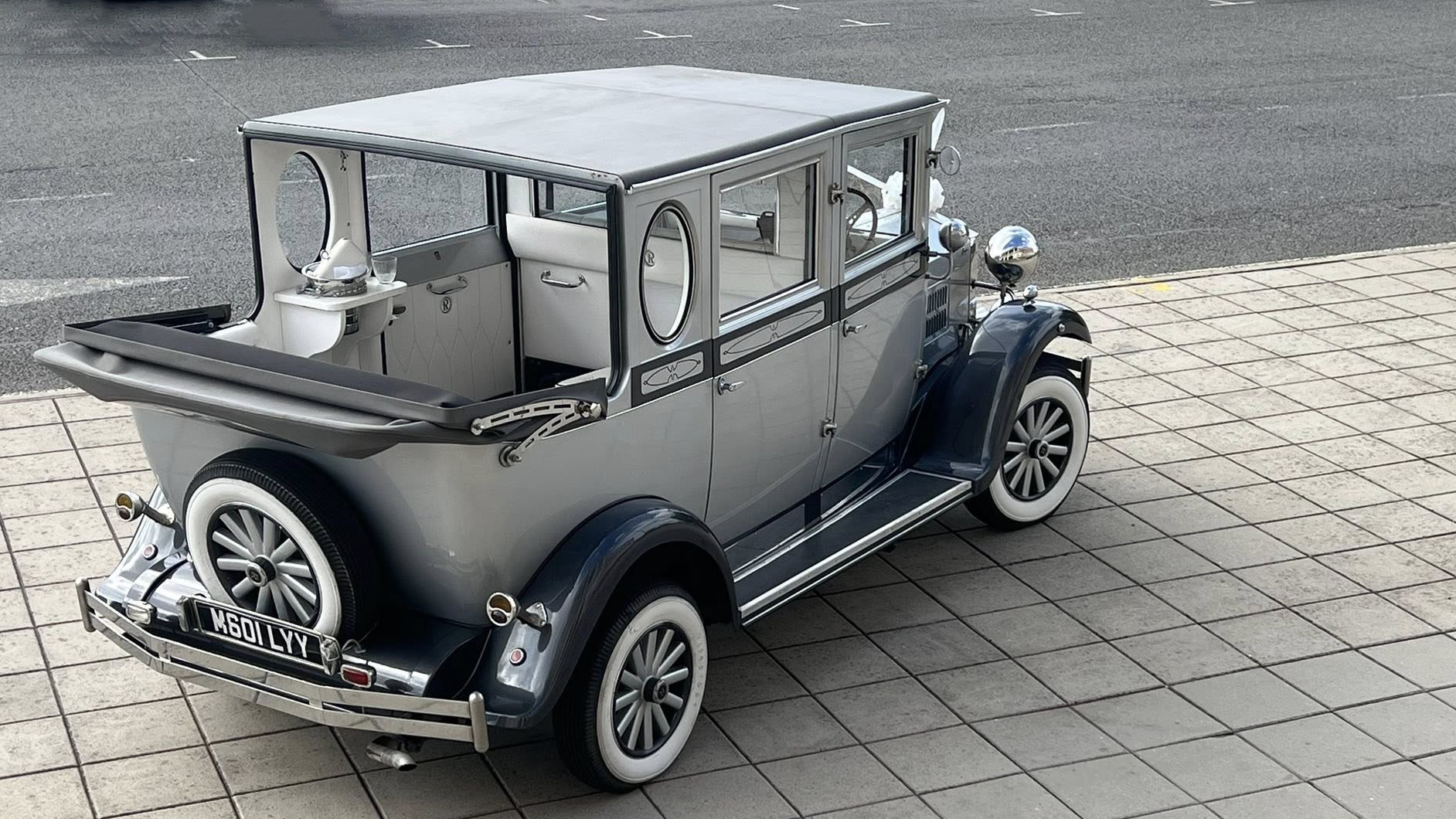 A silver Imperial Landaulette wedding car photographed from an elevated angle with the rear soft-top roof folded down, revealing the cream leather interior. The car sits on a paved driveway with subtle reflections on its grey bodywork and white-spoked wheels visible on both sides.