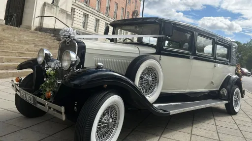 A black and ivory vintage Bramwith-style limousine parked on a stone driveway, featuring side-mounted spare wheels, chrome headlights, and pale pink wedding bows on the front. A large stone building sits in the background.