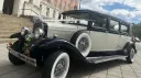 A black and ivory vintage Bramwith-style limousine parked on a stone driveway, featuring side-mounted spare wheels, chrome headlights, and pale pink wedding bows on the front. A large stone building sits in the background.