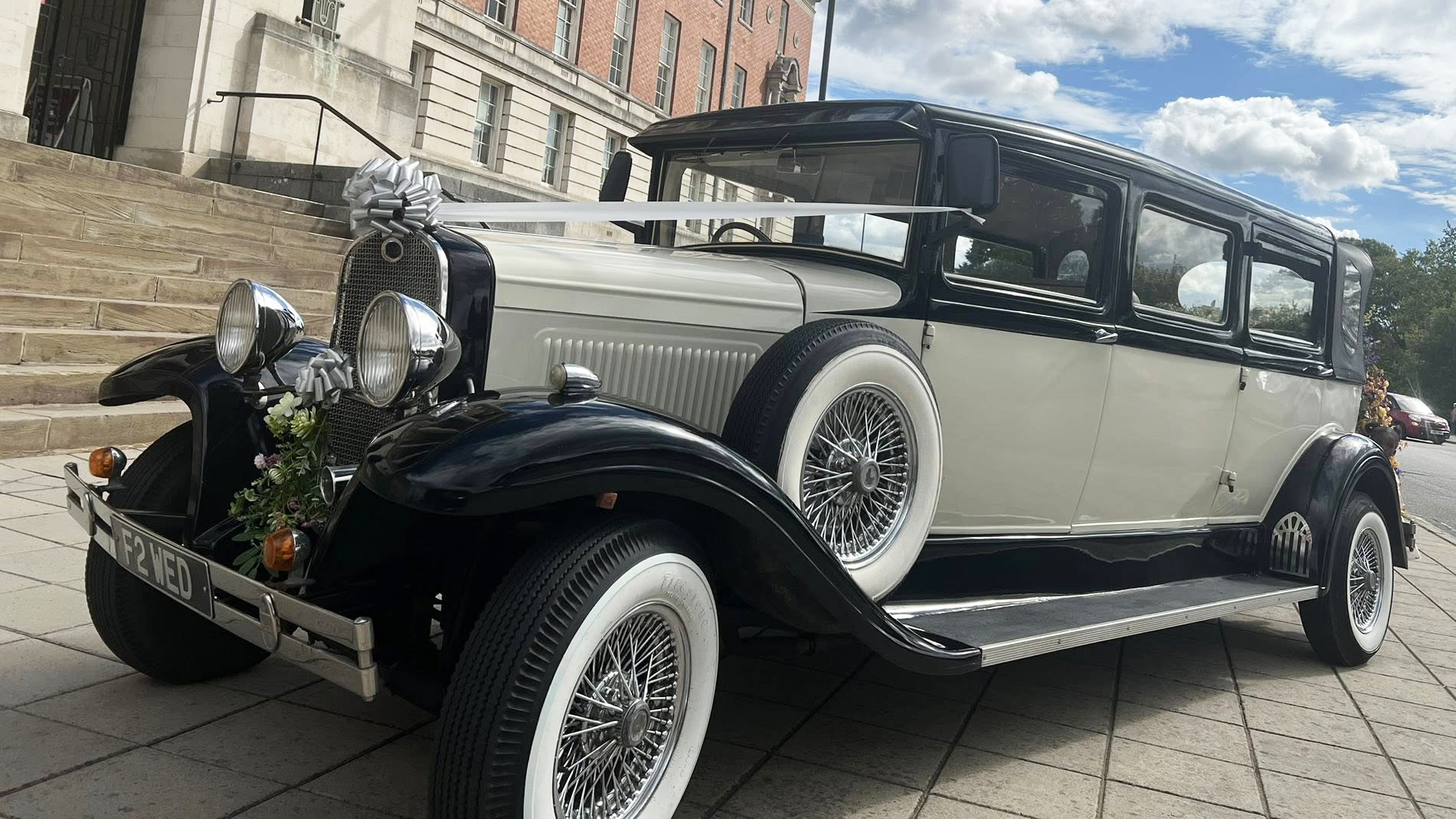 A black and ivory vintage Bramwith-style limousine parked on a stone driveway, featuring side-mounted spare wheels, chrome headlights, and pale pink wedding bows on the front. A large stone building sits in the background.