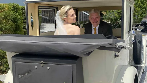 Bride and her father seated inside a cream vintage Viscount wedding car with the soft-top roof folded back, showing the cream leather interior and chrome details, parked in front of a stone building.