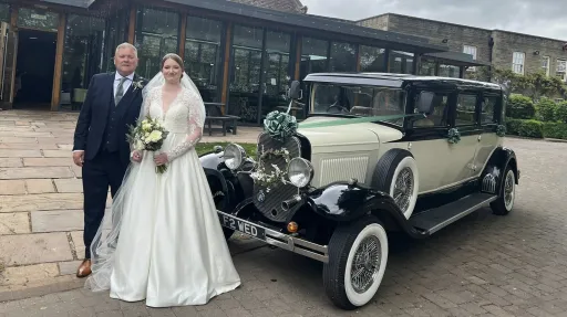 A bride in a white wedding gown holding a bouquet stands beside a black and ivory vintage Bramwith limousine, with the groom in a dark suit by her side. The car has white ribbons, side-mounted spare wheels, and classic chrome detailing. Captured outside a country venue with wooden beams and gardens.