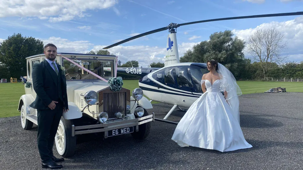 A bride in a white gown and a groom in a dark suit stand beside a cream vintage Imperial wedding car decorated with white ribbons and bows, with side-mounted spare wheels. A helicopter sits on the grass behind them, creating a dramatic backdrop.