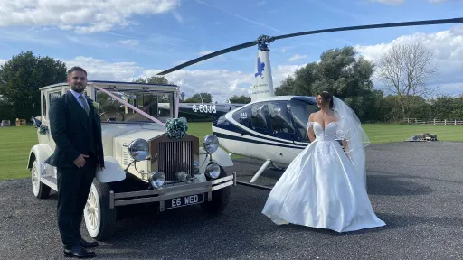 A bride in a white gown and a groom in a dark suit stand beside a cream vintage Imperial wedding car decorated with white ribbons and bows, with side-mounted spare wheels. A helicopter sits on the grass behind them, creating a dramatic backdrop.