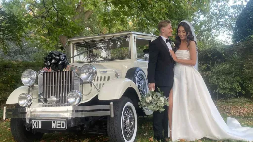 Bride and groom standing beside a cream vintage Viscount wedding car decorated with white ribbon, featuring chrome headlights and a spare wheel on the side, with greenery and trees in the background.