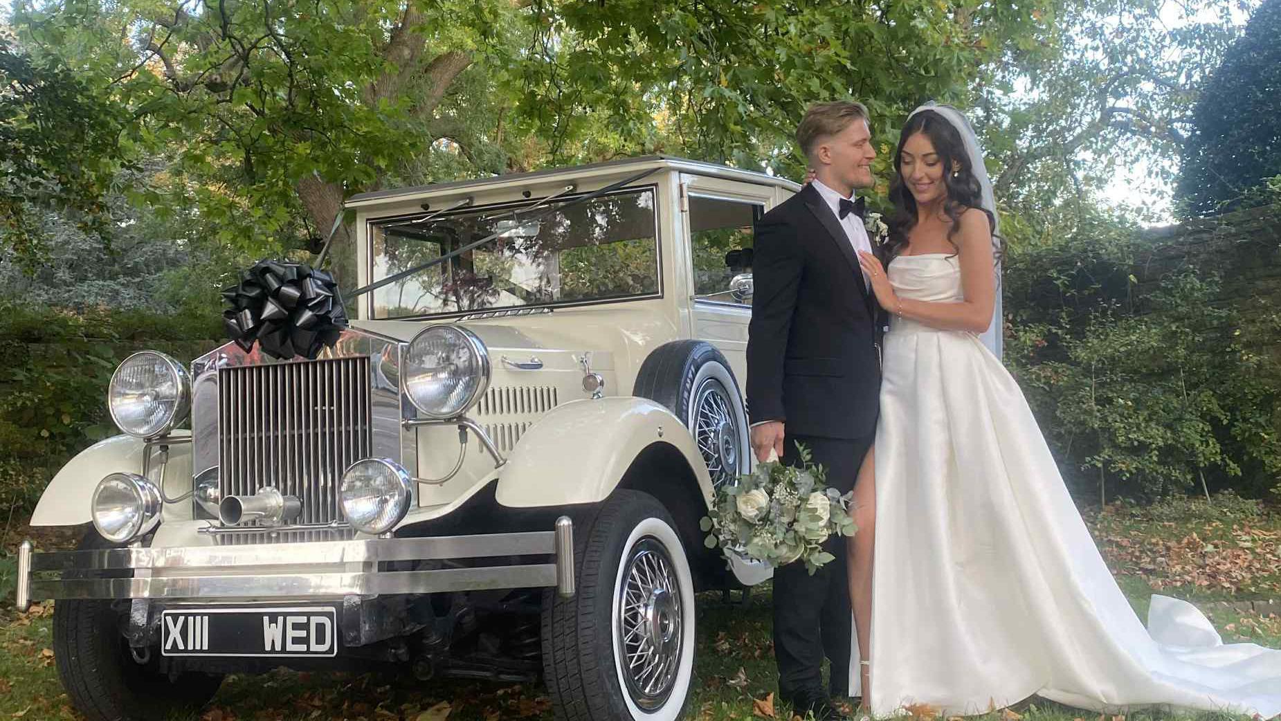 Bride and groom standing beside a cream vintage Viscount wedding car decorated with white ribbon, featuring chrome headlights and a spare wheel on the side, with greenery and trees in the background.