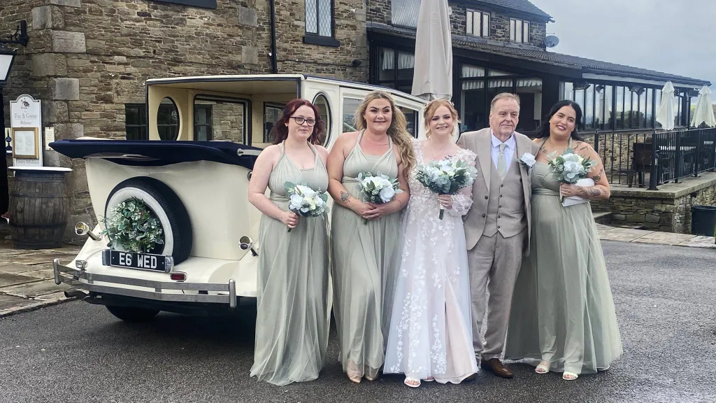 A bride and bridesmaids dressed in soft sage-green dresses stand smiling in front of a cream Imperial vintage wedding car with a blue landaulette roof and white ribbons, parked outside a country hotel with stone walls and large windows.