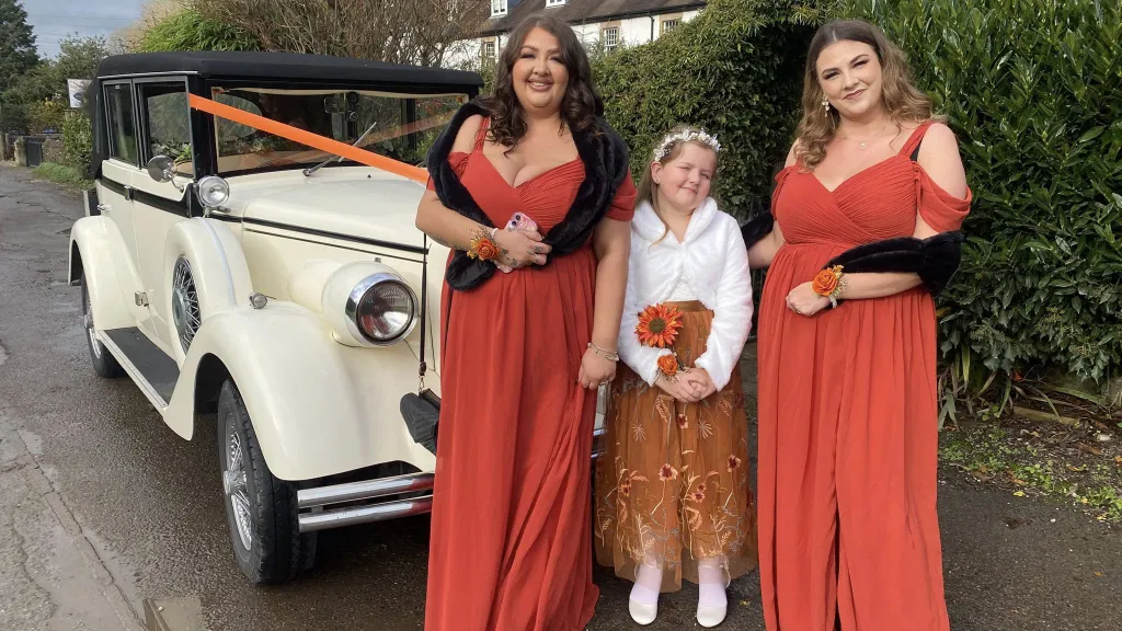 Three bridesmaids dressed in beautiful autumn-coloured gowns standing beside a white and black vintage Regent wedding car, with spare wheel detailing on the side and an orange bow on the roof bar, parked on a driveway with hedges behind them.