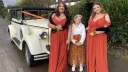 Three bridesmaids dressed in beautiful autumn-coloured gowns standing beside a white and black vintage Regent wedding car, with spare wheel detailing on the side and an orange bow on the roof bar, parked on a driveway with hedges behind them.