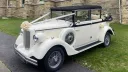 A cream and black vintage Regent convertible wedding car photographed from the front-side angle, showing its classic round headlights, spare side-mounted wheels, and landaulette roof, parked on a grass-bordered driveway.