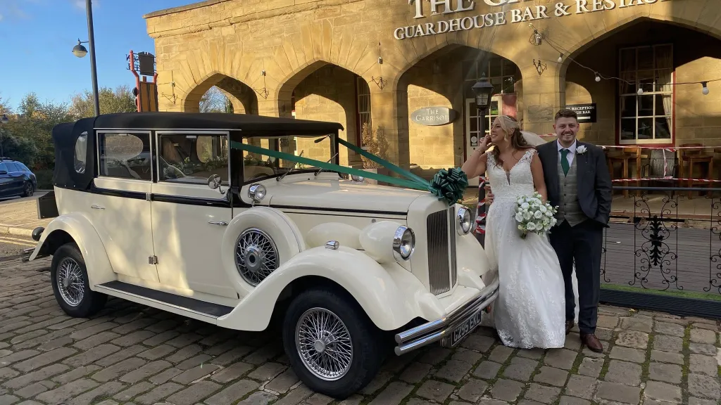 A bride in a white dress holding a bouquet standing beside a groom in a dark suit, both posing with a cream and black vintage Regent wedding car decorated with white ribbons, outside a historic stone building with arched entrances.