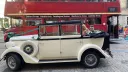 A couple seated inside a cream and black vintage Regent wedding car with the roof open, parked in front of a red London Routemaster bus, with cobblestone ground beneath and city buildings in the background.