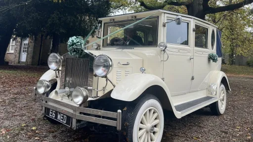 Front-side view of a cream Imperial vintage convertible wedding car with chrome headlamps, exposed spare wheels, and a blue fabric roof. The car is decorated with teal floral ribbons and parked on a leaf-covered driveway in a wooded area in Derbyshire.
