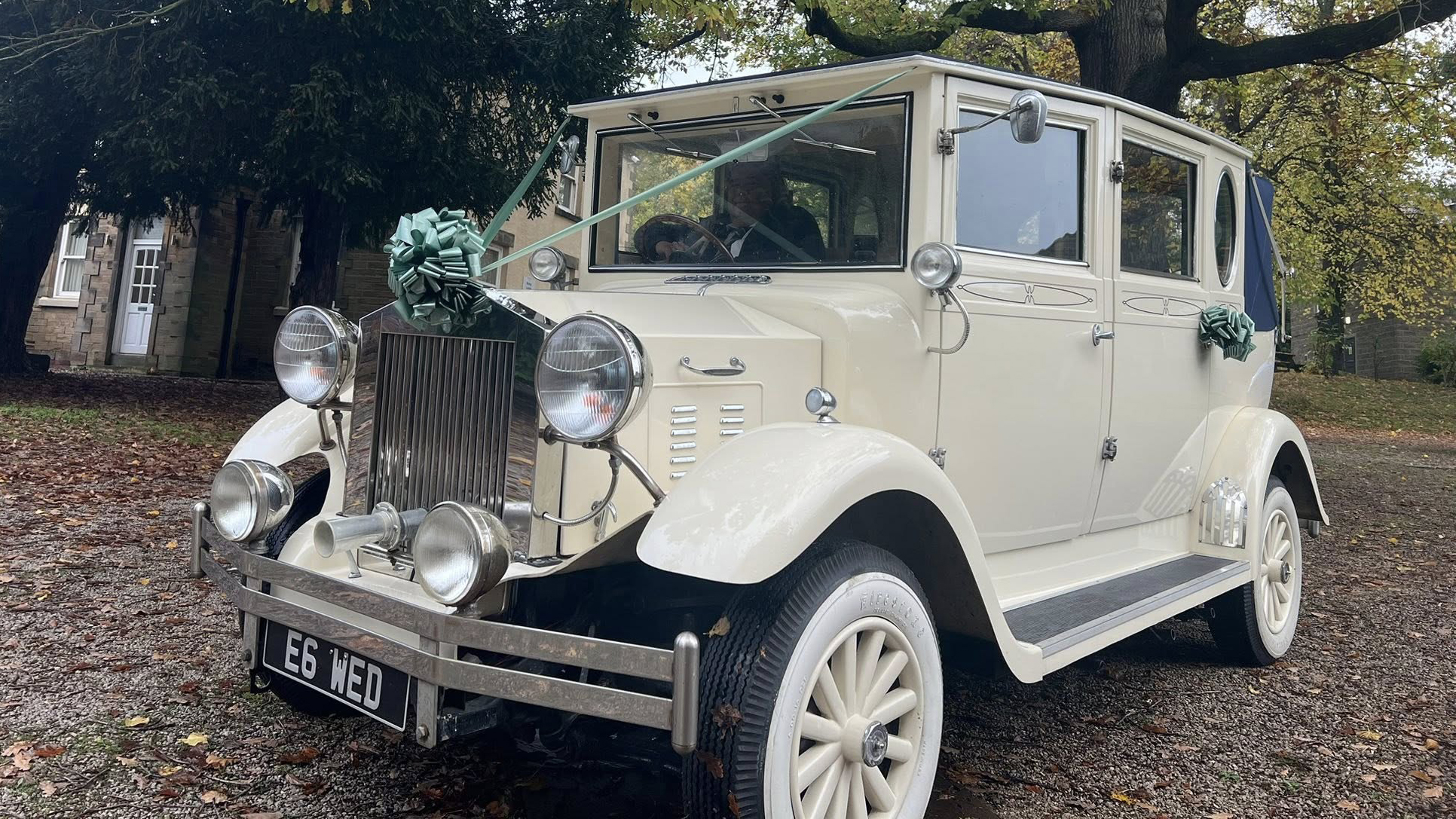 Front-side view of a cream Imperial vintage convertible wedding car with chrome headlamps, exposed spare wheels, and a blue fabric roof. The car is decorated with teal floral ribbons and parked on a leaf-covered driveway in a wooded area in Derbyshire.